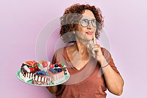 Beautiful middle age mature woman holding cake slices serious face thinking about question with hand on chin, thoughtful about