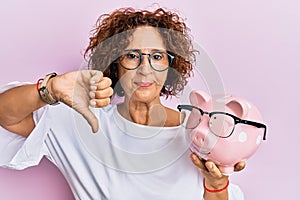 Beautiful middle age mature woman holding piggy bank with glasses with angry face, negative sign showing dislike with thumbs down,