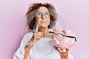 Beautiful middle age mature woman holding piggy bank with glasses serious face thinking about question with hand on chin,