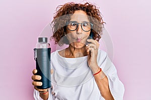 Beautiful middle age mature woman using smartphone holding water bottle making fish face with mouth and squinting eyes, crazy and