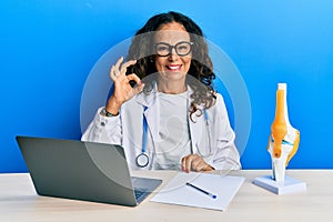 Beautiful middle age woman doctor at orthopedic clinic smiling positive doing ok sign with hand and fingers