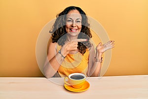 Beautiful middle age woman drinking a cup of coffee amazed and smiling to the camera while presenting with hand and pointing with