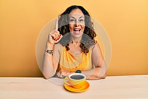 Beautiful middle age woman drinking a cup of coffee pointing finger up with successful idea