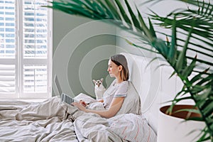 Beautiful middle age woman sitting in bed in morning with laptop surfing Internet. Young woman talking with friend in social media