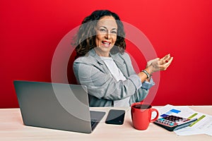 Beautiful middle age woman working at the office drinking a cup of coffee pointing aside with hands open palms showing copy space,