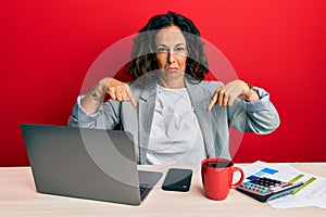 Beautiful middle age woman working at the office drinking a cup of coffee pointing down looking sad and upset, indicating