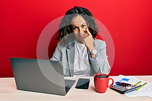 Beautiful middle age woman working at the office drinking a cup of coffee pointing to the eye watching you gesture, suspicious