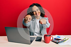 Beautiful middle age woman working at the office drinking a cup of coffee pointing to you and the camera with fingers, smiling