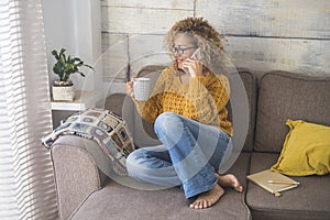 Beautiful middle age woman with yellow sweater do phone call at home sitting on the couch and drinking coffee - relax lifestyle