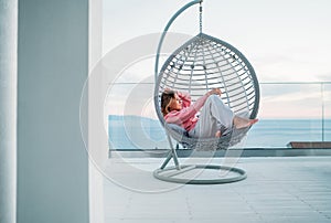Beautiful middle-aged barefoot woman lying in comfortable hanging chair on open house terrace and enjoying the sunset sun rays.
