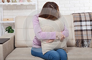 Beautiful middle aged brunette woman holding a pillow and crying on sofa. Home background. Menopause time