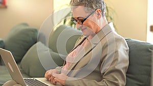 Beautiful middle-aged business lady wearing glasses and a gray business suit is working with a laptop while sitting on