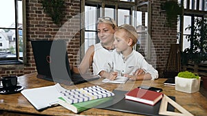 Beautiful middle-aged businesswoman working on laptop when her little lovely grandson something writes in a notebook at