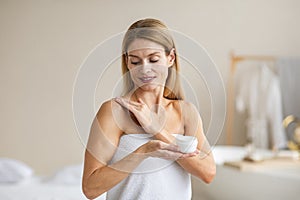 Beautiful middle aged lady wrapped in towel applying body cream on her shoulder after bath in bedroom interior