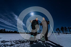 Beautiful middle aged men and women in snowshoes stand in night rare snowy winter forest under full moon light. Night walk Lapland