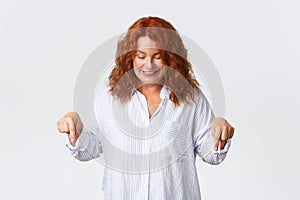 Beautiful middle-aged redhead woman in shirt looking and pointing fingers down with happy, cute smile. Concept of promo