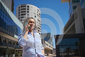 Beautiful middle aged woman in casual clothes, talking on mobile phone, against modern business buildings background