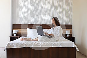 Beautiful middle aged woman dressed in white bathrobe sitting on bed and working with laptop in hotel room