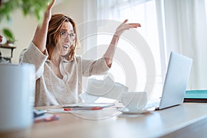 Beautiful middle-aged woman in glasses extremely surprised with new higher taxes watching, throwing bills at laptop in home office