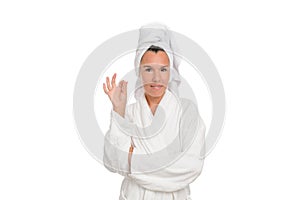 A beautiful middle-aged woman in a white robe and a towel on her head smiles and shows an OK gesture.