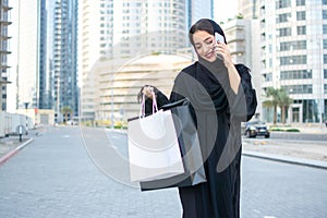 Beautiful middle eastern woman holding shopping bags and talking on cellphone while walking on the street.