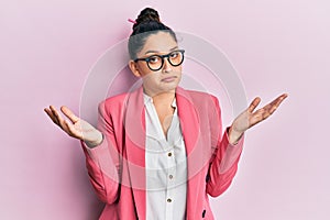 Beautiful middle eastern woman wearing business jacket and glasses clueless and confused expression with arms and hands raised