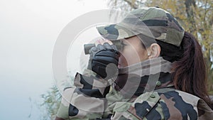 Beautiful military woman in army camouflage looking into binoculars on the river in the fog on an autumn morning