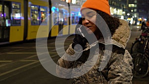 Beautiful mixed race female teenager girl young woman talking at night on cell phone at night with trams by Alexanderplatz Station