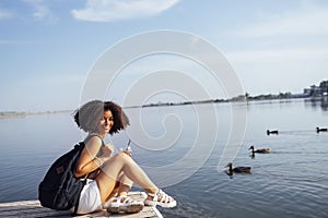 Beautiful mixed race female teenager sits on wooden pier and drinks cocktail in plactic glass