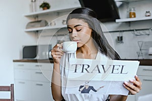 Beautiful mixed race girl in white robe drinking coffee and holding travel newspaper in morning