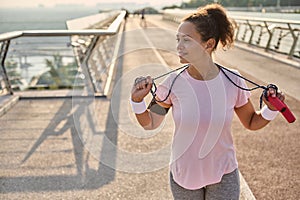Beautiful mixed race woman in sportswear standing on a treadmill on the city bridge with a jumping rope, looking aside while