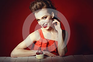 Beautiful model with creative hairstyle and colourful make up sitting at the wooden table with delicious cherry pastry on it