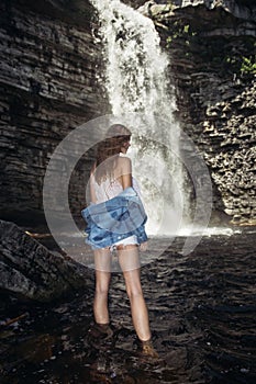 Beautiful model girl with long legs posing near a waterfall wearing jeans jacket.