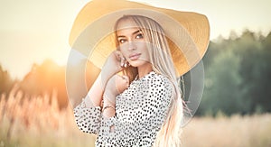 Beautiful model girl posing on a field, enjoying nature outdoors in wide brimmed straw hat. Beauty blonde young woman