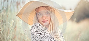 Beautiful model girl posing on a field, enjoying nature outdoors in wide brimmed straw hat. Beauty blonde young woman