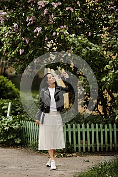 Beautiful model girl in a white midi skirt and black leather jacket posing near a bloomy lilac tree. Young brunette