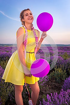A beautiful model with long braids stands in a lavender field. Holds two balloons in his hands. Laughs sticking his tongue out.
