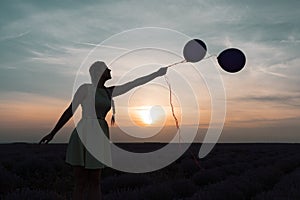 A beautiful model with long braids stands in a lavender field. Holds two balloons in his hands. Silhouette photo. Sunset in the