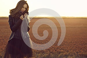 Beautiful model with long hair wearing black veiling dress and stylish leather jacket standing in the deserted field at sunset