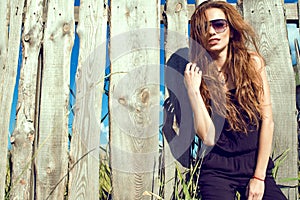 Beautiful model wearing black jumpsuit and trendy aviator sunglasses standing at the old shabby fence made of boards