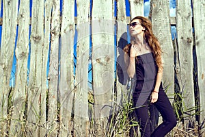 Beautiful model wearing black strapless jumpsuit and trendy sunglasses standing at the old shabby wooden fence photo