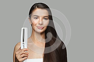 Beautiful model woman holding hair iron and straightening her healthy long dark hair on white background.