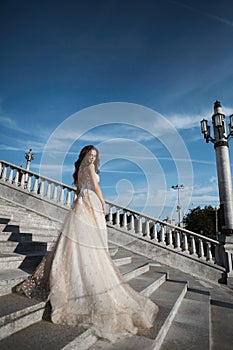 Beautiful model woman with perfect body in luxury wedding dress stands with back on the stairs and posing with blue sky