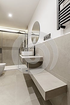 Beautiful modern bathroom with gray tiles, countertop of the same material, circular mirror, shower cabin with glass screens, wall