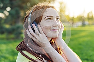 Beautiful modern girl with dreadlocks listening to music with headphones in autumn Sunny Park. Melomania and good mood