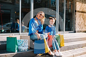 Beautiful mom and her cute little daughter are holding shopping bags, looking at camera and smiling while standing outdoors.