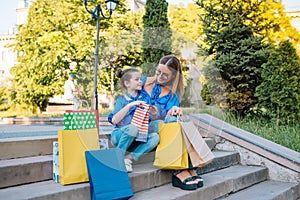 Beautiful mom and her cute little daughter are holding shopping bags, looking at camera and smiling while standing outdoors.