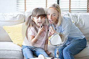 Beautiful mom with her little cute daughter are doing your makeup and having fun. Mom and daughter wear lipstick look at mirror