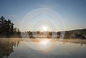 Beautiful morning orange sunrise over lake Sjabatjakjaure with haze mist in Sweden Lapland nature. Mountains, birch trees, spruce