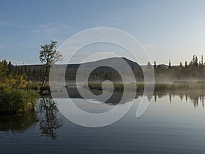 Beautiful morning over lake Sjabatjakjaure with haze mist in Sweden Lapland nature. Mountains, birch trees, spruce forest, rock